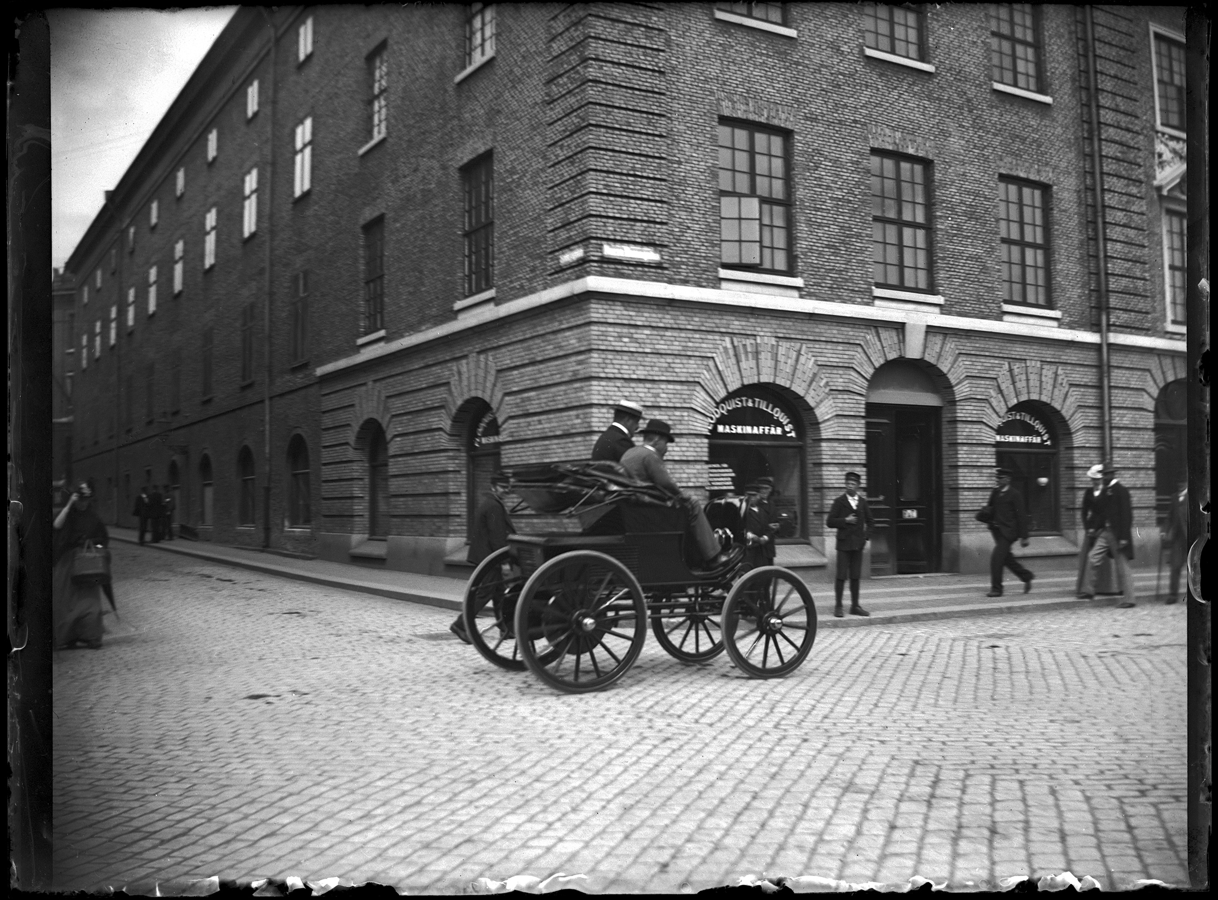Två män åker i en äldre bil år 1900 medan människor står utmed gatan och kollar på.